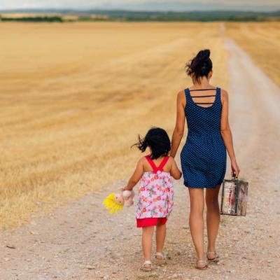 Eine Frau und ein Kind gehen Hand in Hand einen Feldweg zwischen gelben Feldern entlang.