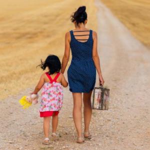 Eine Frau mit einem Kinder an der Hand gehen mit dem Rücken zum Betarchter einen sandigen Weg entlang.